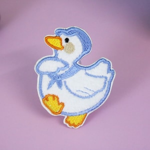 May include: A blue and white embroidered iron-on patch of a duck with yellow feet and beak.