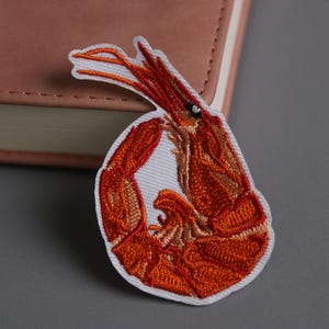 May include: Embroidered patch depicting an orange and brown shrimp with a white border. The patch is on a grey surface beside a closed pink notebook. The shrimp design has detailed stitching.