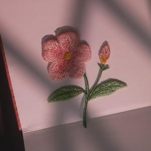 May include: Embroidered pink flower with a yellow centre and a bud, set against a soft pink background. The flower has detailed stitching, green leaves, and a green stem. The image is well-lit, highlighting the texture of the embroidery.