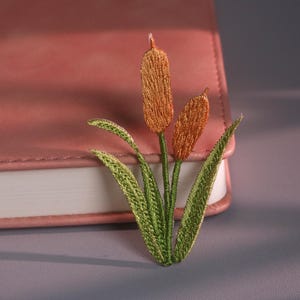 Op de afbeelding: Geborduurde lisdodde-patch met twee bruine zaadkoppen en groene bladeren. De patch is geplaatst voor een roze leren notitieboek op een grijze ondergrond. Het lisdodde-ontwerp is gedetailleerd.