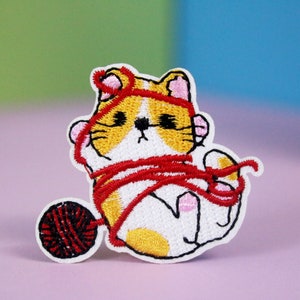 May include: Embroidered iron-on patch of a white cat with orange and yellow markings tangled in red yarn. The cat is looking down with a sad expression. The cat is sitting next to a ball of red yarn.