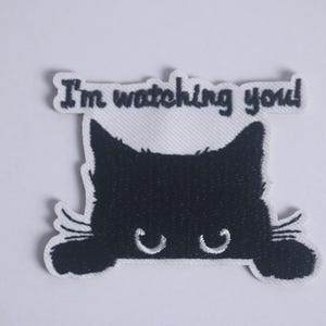 May include: A black cat patch with the text "I'm watching you!" in black lettering. The cat's silhouette is black with white accents, including the eyes and whiskers. The patch is on a white background.
