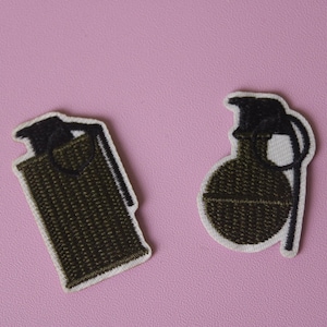 May include: Two embroidered patches depicting hand grenades. One is a green and black grenade with a pin, the other is a green and black grenade with a handle.