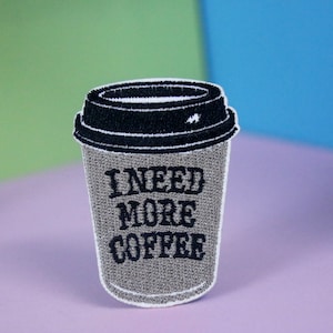 May include: A grey embroidered iron-on patch of a coffee cup with the text "I NEED MORE COFFEE" in black.