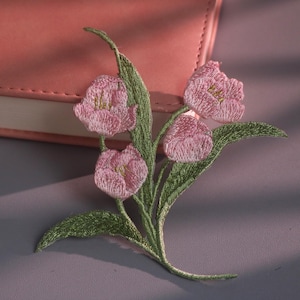 Può includere: Una toppa ricamata raffigurante tre fiori rosa chiaro simili a tulipani con centri gialli e steli e foglie verdi dettagliati. Il ricamo utilizza filo testurizzato per creare profondità e dimensione sui petali e sul fogliame del disegno.