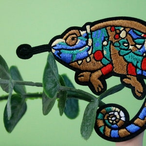May include: A colorful embroidered patch of a chameleon with a blue, green, red, and brown design. The chameleon is curled up with its tail wrapped around its body.
