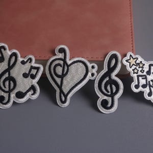 May include: Four embroidered patches with musical themes. The patches feature black treble clefs, musical notes, and stars on a white background with a white border. The patches are arranged on a gray surface with a pink notebook in the background.