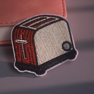 May include: An embroidered patch shaped like a toaster with a red side, a light gray front, and a black base. The design features a vertical silver lever on the side, a circular gray dial on the front, and a white border outlining the entire shape.
