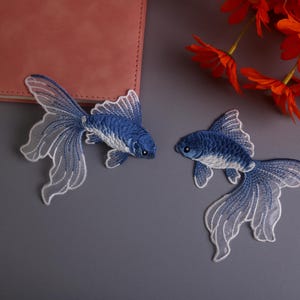 Może przedstawiać: Dwie haftowane naszywki w kształcie złotych rybek. Ryby są niebieskie z białymi akcentami i delikatnymi, koronkowymi płetwami. Leżą na szarym tle z różowym notatnikiem i pomarańczowymi kwiatami.