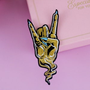 Puede incluir: Un parche de hierro bordado en oro que representa una mano de esqueleto haciendo el gesto de rock and roll. La mano est&aacute; delineada en negro y tiene u&ntilde;as azules.