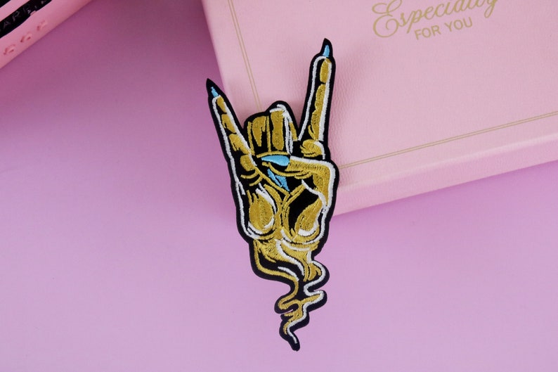 Puede incluir: Un parche de hierro bordado en oro que representa una mano de esqueleto haciendo el gesto de rock and roll. La mano est&aacute; delineada en negro y tiene u&ntilde;as azules.