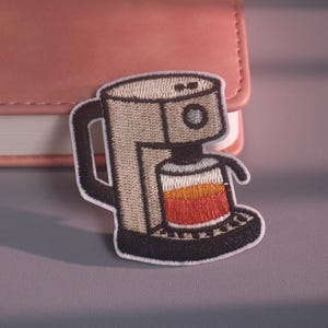 以下が含まれることがあります： コーヒーメーカーの形をした刺繍入りのアイロン接着ワッペン。本体はベージュで、ハンドルとベースは黒色です。ガラス製のカラフェには、上部が黄色、中央がオレンジ、下部が赤色の層になった液体が入っています。