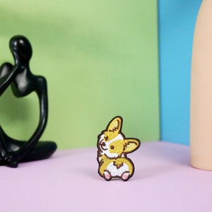 Może przedstawiać: Naszywka haftowana przedstawiająca kresk&oacute;wkowego psa rasy corgi z uśmiechem. Naszywka jest brązowa i biała i znajduje się na r&oacute;żowym tle.