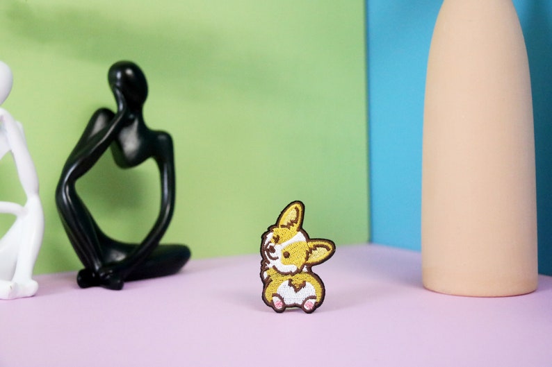 Może przedstawiać: Naszywka haftowana przedstawiająca kresk&oacute;wkowego psa rasy corgi z uśmiechem. Naszywka jest brązowa i biała i znajduje się na r&oacute;żowym tle.