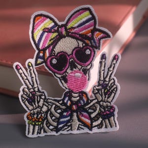 May include: An embroidered patch featuring a skeleton wearing a multi-colored striped bow, heart-shaped sunglasses, and a matching neck scarf. The skeleton is blowing a pink bubble with gum and holding up both hands in a peace sign gesture.