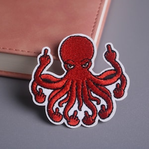 Puede incluir: Parche de pulpo rojo bordado con borde blanco. El pulpo tiene una expresión enfadada y muestra el dedo corazón con cada tentáculo. El parche está sobre una superficie gris junto a un libro rosa.