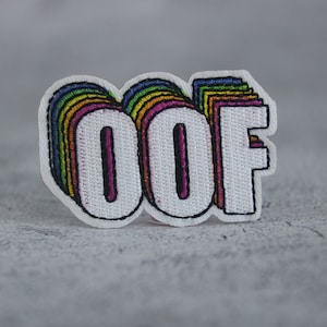 May include: A white embroidered patch with the word "OOF" in rainbow colors. The letters are outlined in black and have a 3D effect.