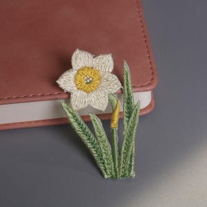 Puede incluir: Parche de narciso bordado con pétalos blancos y centro amarillo, hojas verdes y tallo amarillo. El parche floral está colocado sobre un cuaderno de cuero rosa, sobre un fondo gris. Un artículo decorativo.