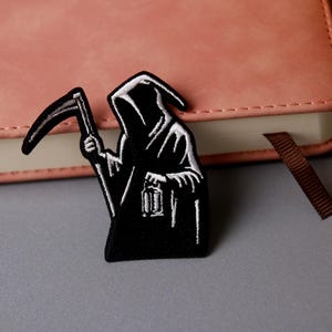 May include: Black and white embroidered patch of the Grim Reaper holding a scythe and a lantern. The patch is on a pink leather notebook.