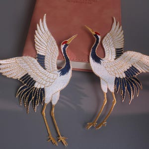 May include: Two embroidered crane patches featuring white bodies, dark blue wing and tail feathers, and red accents on the heads. The birds are depicted in flight with outstretched wings and long, thin yellow legs, showcasing detailed stitching throughout.