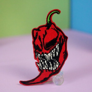 May include: A red chili pepper-shaped patch with a scary face design. The patch has a black outline and a green stem. The pepper's face has sharp white teeth and a sinister expression, ideal for jackets, bags, or other accessories.