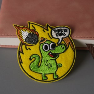 May include: A yellow embroidered patch with a green dinosaur character wearing a hat. The dinosaur is standing in front of a yellow flame with a black meteor. The patch has the text "This is fine!" in a speech bubble.