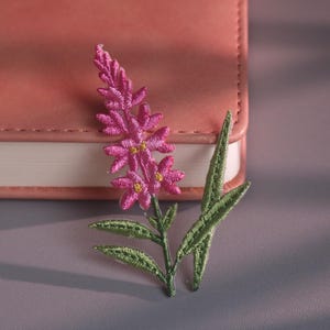 Könnte beinhalten: Eine rosa gestickte Blume mit grünen Blättern liegt auf einer grauen Oberfläche neben einem rosa Notizbuch. Die Blume hat kleine gelbe Zentren und ist mit aufwendigen Stichen versehen. Das Notizbuch hat einen genähten Rand und weiße Seiten.