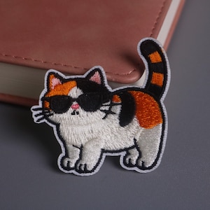 May include: Embroidered patch featuring a cool calico cat wearing sunglasses. The cat has white, orange, and black fur, with a black and orange striped tail. The patch is on a gray surface.