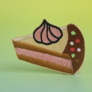 Op de afbeelding: Een geborduurde patch van een stuk cake met een roze swirl van frosting erop, in bruin, roze en wit.