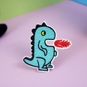 Puede incluir: Un parche bordado azul de un dragón de dibujos animados que escupe fuego. El dragón está contorneado en negro y tiene un ojo amarillo.