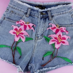 May include: A pair of denim shorts with two embroidered pink lily flowers on each leg. The flowers have green leaves and brown stems.