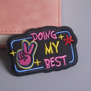 Può includere: Toppa ricamata con le parole "DOING MY BEST" in rosa e giallo, insieme a un gesto della mano del segno della pace. La toppa ha uno sfondo nero con un bordo blu e una stella rossa.