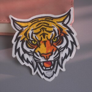 May include: An embroidered patch featuring the head of a tiger with yellow, orange, and white fur, black stripes, and piercing yellow eyes. The tiger is depicted with an open mouth showing teeth, outlined in thick black thread on a white background.