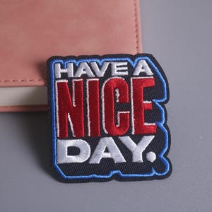 May include: Embroidered patch with the words "HAVE A NICE DAY" in red, white, and blue. The patch has a black background and a blue border. The patch is placed on a gray surface next to a pink notebook.