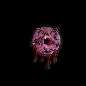 Könnte beinhalten: Gestickter Aufnäher mit einem rosa Augapfel mit roten Adern und heruntertropfendem Blut. Das Auge hat eine schwarze Pupille und einen weißen Lichtreflex. Geeignet für Kleidung oder Accessoires.