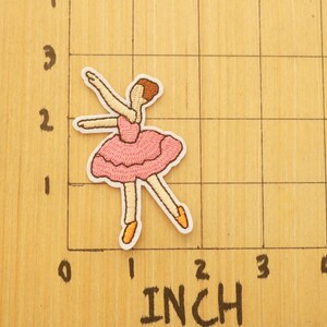 May include: Embroidered patch of a ballerina in a pink tutu and ballet shoes. The patch is approximately 2 inches tall. The ballerina is in a graceful pose with arms extended. The patch has a white border.
