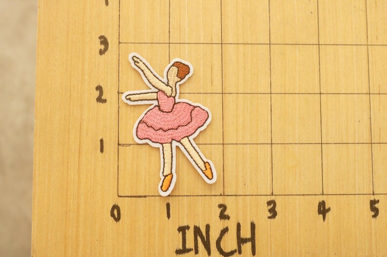 May include: Embroidered patch of a ballerina in a pink tutu and ballet shoes. The patch is approximately 2 inches tall. The ballerina is in a graceful pose with arms extended. The patch has a white border.