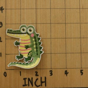 May include: Embroidered alligator patch, approximately 2 inches tall, featuring a green body, yellow striped belly, and pink cheek accents. The patch is on a wooden surface with inch markings.