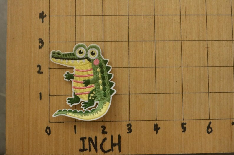 May include: Embroidered alligator patch, approximately 2 inches tall, featuring a green body, yellow striped belly, and pink cheek accents. The patch is on a wooden surface with inch markings.