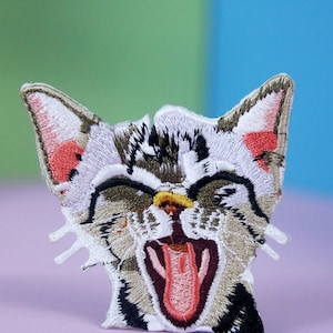 以下が含まれることがあります： 口を開けた猫の刺繍パッチ。ピンクの舌と黄色の歯が見える。グレー、茶色、白の色合いで詳細にデザインされており、耳には赤いアクセントがある。猫はあくびをしているか、咆哮しているように見える。