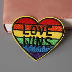 May include: A rainbow heart-shaped patch with the text "LOVE WINS" embroidered in black thread.
