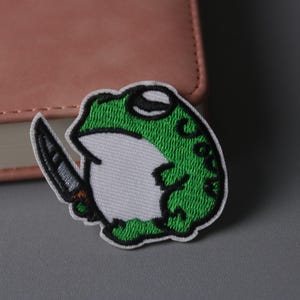 Może przedstawiać: Naszywka z haftowanym zielonym żabą trzymającym nóż. Żaba jest biała z zielonymi plamkami i czarnym okiem. Nóż jest srebrny z brązową rękojeścią.