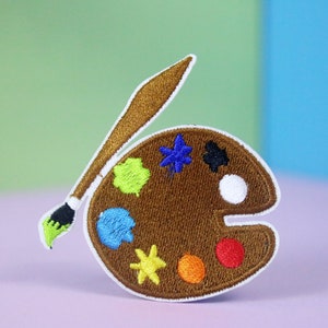 Può includere: Una tavolozza da artista in feltro marrone con un pennello. La tavolozza ha otto pozzetti di vernice di diversi colori. I colori della vernice sono verde, blu, bianco, nero, viola, giallo, arancione e rosso.