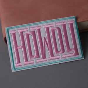 Puede incluir: Un parche bordado en rosa y blanco con la palabra "HOWDY" en rosa claro. El parche tiene un borde turquesa y blanco.