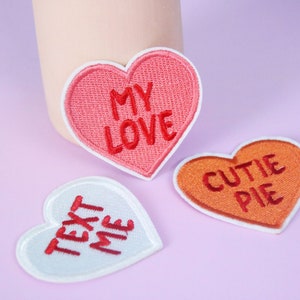 May include: Three heart-shaped embroidered patches with different messages. The first patch is pink with white stitching and says "MY LOVE". The second patch is orange with white stitching and says "CUTIE PIE". The third patch is white with red stitching and says "TEXT ME".