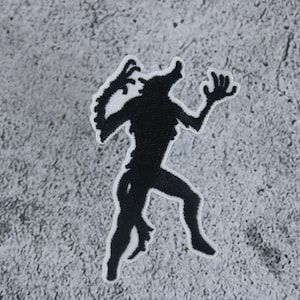 Peut inclure: Patch brodé noir représentant la silhouette d'un loup-garou avec un contour blanc. Le loup-garou est debout sur ses pattes arrière, les bras levés.