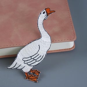 Könnte beinhalten: Gestickter Aufnäher mit einer weißen Gans mit schwarzen Details an Flügeln und Füßen. Die Gans hat einen orangefarbenen Schnabel und Füße sowie einen kleinen orangefarbenen Fleck auf dem Kopf. Der Aufnäher liegt auf einem rosa Notizbuch.