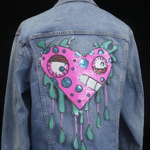 May include: A blue denim jacket with a pink heart-shaped patch on the back. The patch has a cartoon face with blue eyes and a green dripping design.