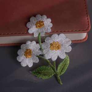 May include: Embroidered daisy patch featuring three white flowers with yellow centers and green stems and leaves. The patch is placed on a brown leather notebook, set against a gray background. This floral patch is perfect for adding a touch of nature to clothing or accessories.