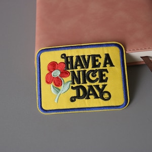 May include: Yellow embroidered patch with blue trim featuring a red flower and the text "HAVE A NICE DAY" with a smiley face.